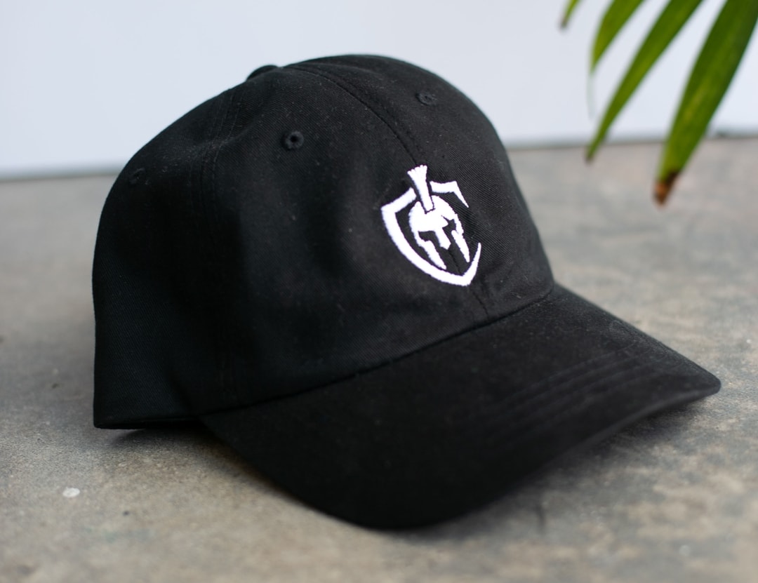 A black dad hat with white embroidery sits on a concrete floor.
