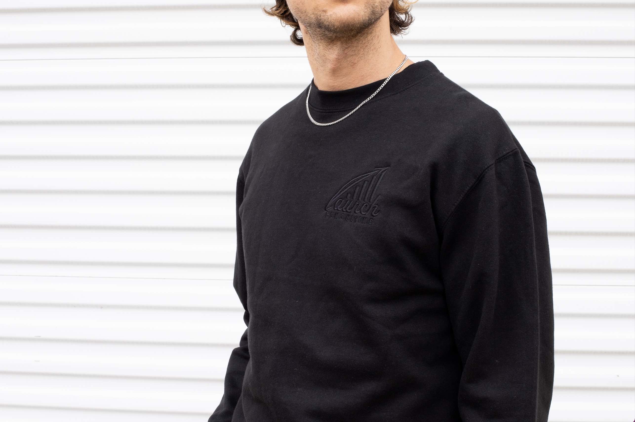 Man wears a black sweatshirt with black embroidered design.